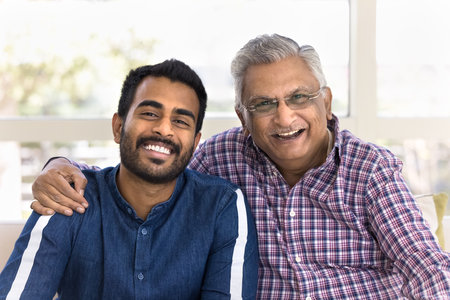 Cheerful elderly Indian father hugging young adult son, touching shoulder with support, parents pride, looking at camera with toothy smiles, laughing, having fun, posing for head shot portraitの写真素材