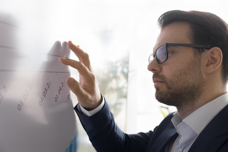 Close up skilled serious businessman analyze infographic data, stand near whiteboard with chart and diagrams, make analysis, engrossed in strategy development working alone. Trading, banking, researchの写真素材