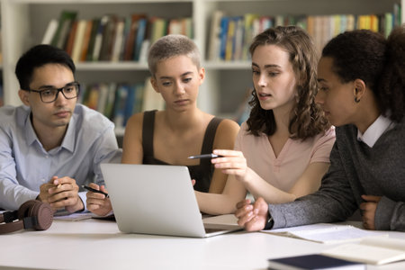 Four focused multiethnic students do homework together sit in library, discuss online task use laptop, prepare for exams, search information on internet, share ideas, work on group project. Educationの写真素材