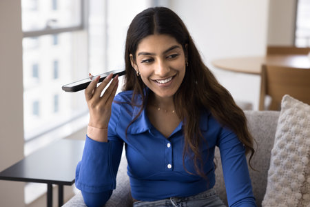 Cheerful young Indian woman holding smartphone at ear, listening to voice message on telephone,...