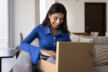 Happy young Indian consumer woman opening logistic cardboard box with plastic wrap inside, receiving...