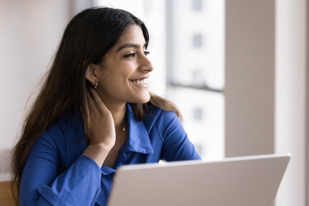 Happy Indian freelance worker girl sitting at laptop, touching neck in deep positive thoughts, looking away, smiling, dreaming, thinking on Internet communication technologyの写真素材