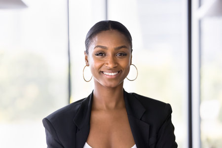 Happy attractive African American business professional woman in formal jacket looking at camera with toothy smile, posing indoors. Positive successful young entrepreneur, manager lady portraitの写真素材