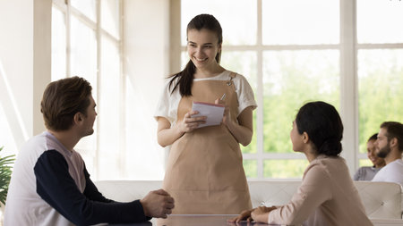 Pretty young waitress wearing uniform apron, serving diverse couple in restaurant, taking order, making notes, smiling, listening to customers, telling about menu, mealの写真素材