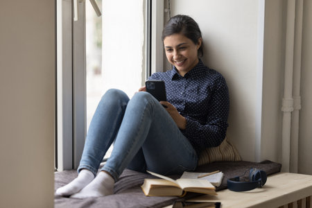 Happy beautiful Indian student woman resting on windowsill with mobile phone, books for studying,...