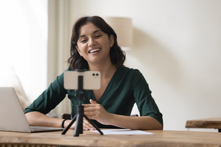 Cheerful pretty professional influencer woman shooting content for blog on smartphone web camera,...