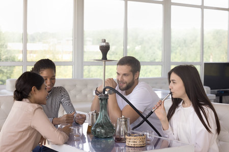 Diverse group of young millennial girls and guy enjoying leisure in hookah lounge, social...