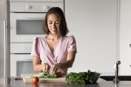 Positive attractive young housewife woman slicing fresh vegetables for salad, cutting green leaves on chopping board, enjoying culinary activity, healthy cooking from organic ingredientsの写真素材
