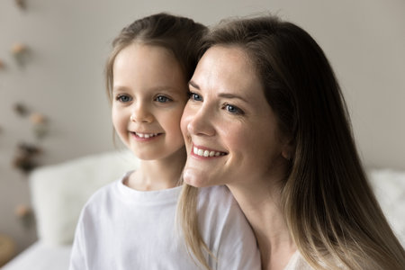 Close up portrait of pretty young mother and little daughter, smiling looking away in morning at home. Family bond and ties, happy woman enjoy motherhood, spend time with adorable preschooler childの写真素材