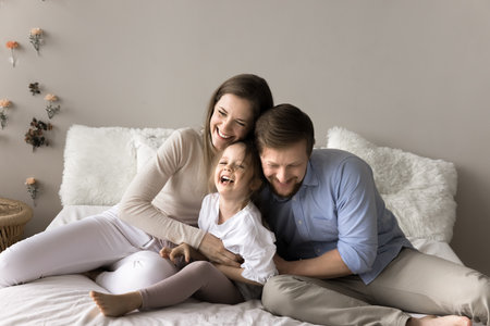 Young laughing parents spend time with preschooler daughter, sitting together on the bed, tickling adorable cheery child, enjoy funny active playtime at home. Family pastime, ties and happy parenthoodの写真素材