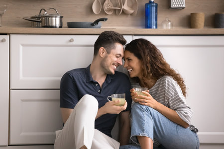 Hispanic couple sit on warm floor with mugs in cozy kitchen, enjoy romantic dating, house-warming, pleasant talk and beverage, spend relocation day to new own house. Cohabitation, relationships, loveの写真素材