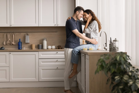 Young married couple sharing tender moment in modern kitchen. Pretty woman sitting on counter while her husband man stands close to her, gently hugging and looking into her eyes with love and warmthの写真素材