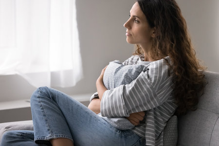 Pensive single woman sits on sofa, gazing out of window in deep contemplation, suffers from solitude, emotional distress, coping with personal troubles and break up, goes through difficult life periodの写真素材