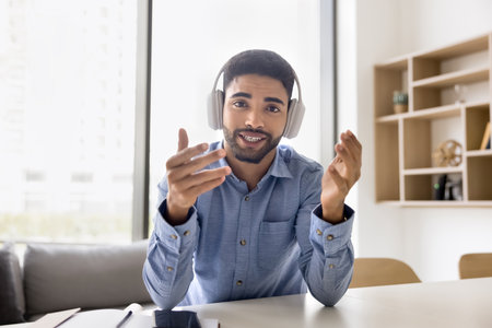 Positive handsome student man in wireless headphones talking on video call, speaking at camera, mobbing hands, smiling. Young Arab blogger, influencer broadcasting on Internet. Screen shot portraitの写真素材