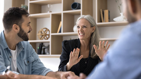 Cheerful senior business leader woman talking to team of younger employees, speaking with toothy...