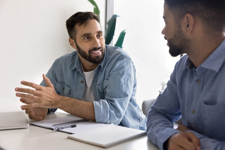 Positive handsome businessman talking to male colleague at workplace table. Diverse coworkers, two employees meeting for job discussion, cooperation on project, startupの写真素材