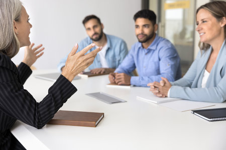 Confident older business leader woman talking to diverse team of younger colleagues at meeting, sitting at large table, giving seminar, training, coaching. Female boss speaking to co-workersの写真素材