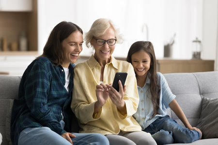 Happy grandmother using smartphone, sitting on couch with cheerful granddaughter kid and her mom, showing online application, Internet content, media service to younger generationsの写真素材