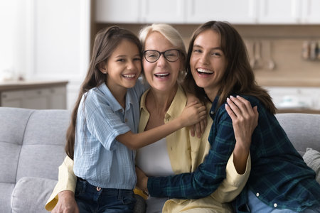 Cheerful kid girl and young mom embracing happy grandma, sitting close together on home couch,...