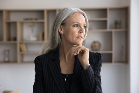 Thoughtful mature businesswoman looking away in office, touching chin, in deep thoughts, making decision, feeling doubts, thinking on questions, planning business projectの写真素材