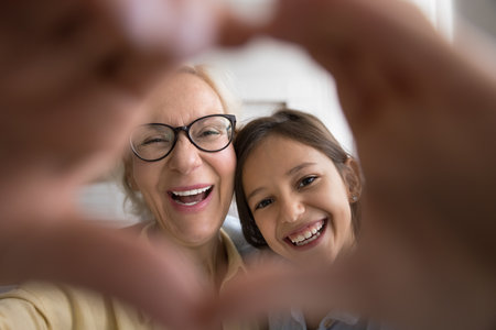 Positive excited granny and happy grandkid girl showing hand heart, gesture of friendship, love, looking at camera through finger frame with toothy smile, posing for family portraitの写真素材