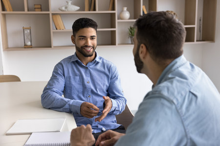 Positive handsome young Arab professional man talking to male colleague at workplace table, speaking, smiling, discussing cooperation, teamwork, startup, creative task, giving consultationの写真素材