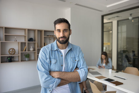 Positive handsome Hispanic project leader man posing in office meeting room space, looking at camera with arms crossed, standing for professional portrait with colleagues working in backgroundの写真素材