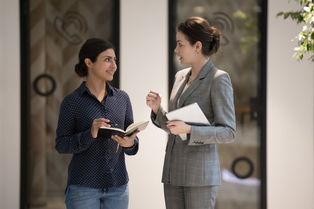 Businesswomen stand in office hallway and talking, boss gives task to Indian secretary at workday in modern workspace. Personal assistant or intern, listens to female chief. Mentoring, apprenticeshipの写真素材