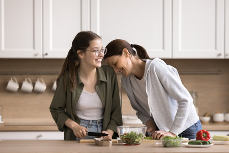 Happy mother and teen daughter cooking in kitchen together, prepare fresh vegetarian salad for dinner, chopping vegetable, communicate, joking, laugh, enjoy culinary, keep diet, lead healthy lifestyleの写真素材