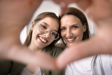 Close up faces of cheerful mother and pretty teen daughter looking at camera, make heart sign with hands, show symbol of love and affection, enjoy tender moment, demonstrate grateful, support and careの写真素材