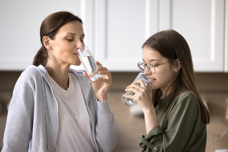 Positive woman and teenage girl stand in modern kitchen, holding glasses, drink natural mineral water, lead healthy life habit, follow diet, caring for health, satisfy thirst. Healthcare, lifestyleの写真素材