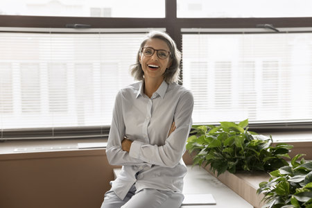 Laughing middle-aged professional businesswoman with short grey hair and eyeglasses posing for camera at modern workplace. Portrait of mature business consultant, project or marketing manager, careerの写真素材