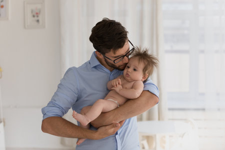 Young loving daddy standing in cozy warm domestic room hugs and kisses gently with love his cute...