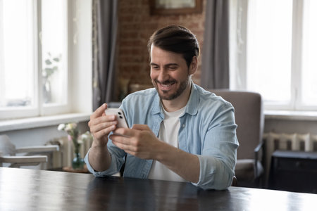Cheerful millennial smartphone user man talking on video call, typing message on mobile phone, using online app, Internet service on wireless gadget at work table, smiling, laughingの写真素材