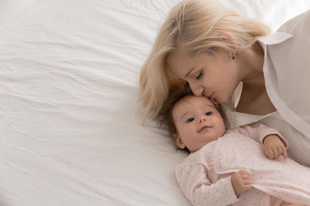 Young loving mother kisses on forehead her lovely newborn, lying together on bed, close up. Mommy...