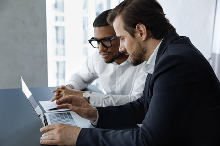 Two multiracial colleagues discuss on-line project, work on presentation seated at desk in office. Diverse businessmen, business partners cooperate during meeting use corporate software, tech conceptの写真素材