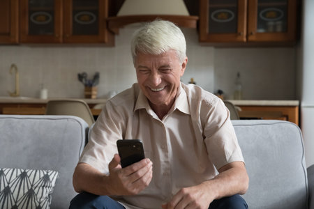 Modern, wireless tech usage. Single older man sits on sofa laughs staring at cellphone screen, watch funny videos content, enjoys new mobile application feels overjoyed spend time at home on internetの写真素材