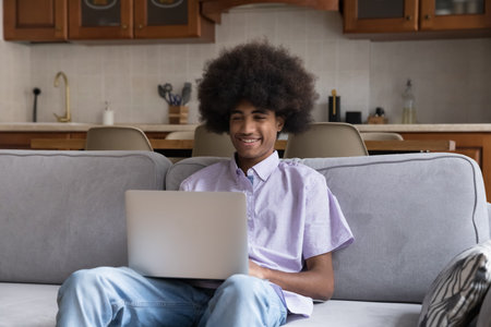 Handsome smiling teenager guy sit on sofa with laptop, watching on-line movie, enjoy videos or vlog on internet, studying online feels satisfied spend weekend at home using modern wireless technologyの写真素材