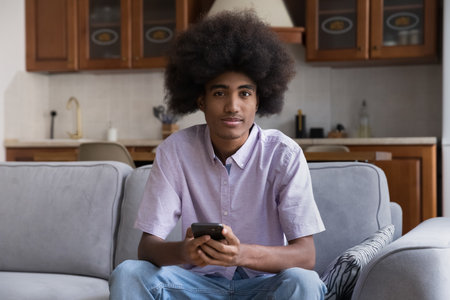 African teenager guy looking at camera seated on sofa with smartphone. Young generation and modern wireless technology overuse, everyday usage of cellphone for communication and leisure on internetの写真素材