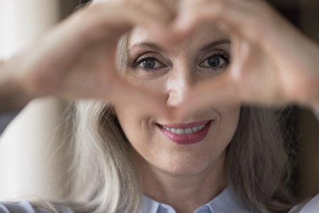 Happy cheerful mature lady looking at camera through hand heart at eyes, upper face. Cheerful senior lady showing gesture if kindness, promoting healthcare, Valentines day. Eyesight, vision conceptの写真素材