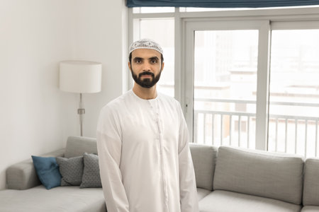 Serious handsome Middle Eastern man in Muslim cloth and headwear home portrait. Arabian model in white kandura and skullcap standing in modern apartment, looking at camera, posingの写真素材