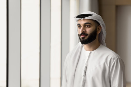 Positive handsome young Arabic businessman wearing white keffiyeh, standing at office window, looking at copy space away, thinking on company leadership, business success, investmentの写真素材