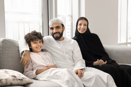 Positive Arabian mom and dad relaxing on comfortable home couch, embracing cheerful little son boy, looking at camera, smiling, laughing, having fun, enjoying family leisure, close relationshipの写真素材