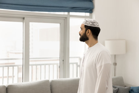Thoughtful serious Middle Eastern man in white Arabic cloth and headwear posing in home apartment, standing in living room, looking at window away, thinking, planning futureの写真素材