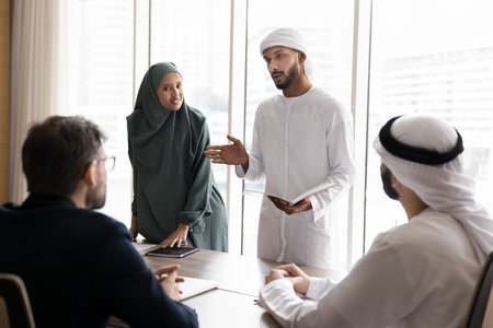 Couple of young Arabian managers in Muslim clothes presenting project to European business partners, speaking to colleagues sitting at meeting table, discussing company selling, investmentの写真素材