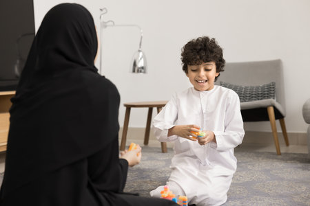 Positive adorable Arabic son boy and mother in Muslim cloth playing with constructor together. Kid...
