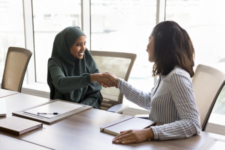 Positive young Arab businesswoman in hijab and female Latin project partner shaking hands at meeting...