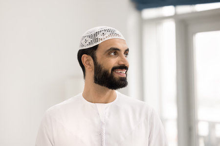 Positive pensive handsome Arabian man in white Muslim cloth and headwear posing in home apartment with white background, looking away, smiling, thinking, daydreaming, planning optimistic futureの写真素材