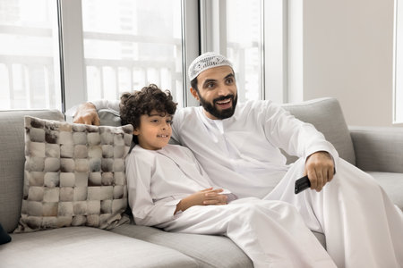 Cheerful Muslim father and positive son boy watching TV show, movie, series, video together, setting channel with remote control, enjoying leisure on home couch with online media serviceの写真素材