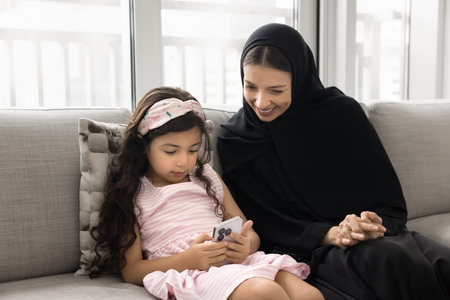 Happy Muslim mom watching little daughter kid using mobile phone, playing game for children on...